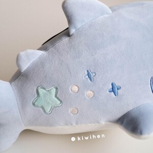 May include: A light blue plush whale with embroidered details, including a green star, white dots, and blue crosses. The whale has a white belly and a fin on its back. The text "kiwihen" is embroidered on the whale's belly.