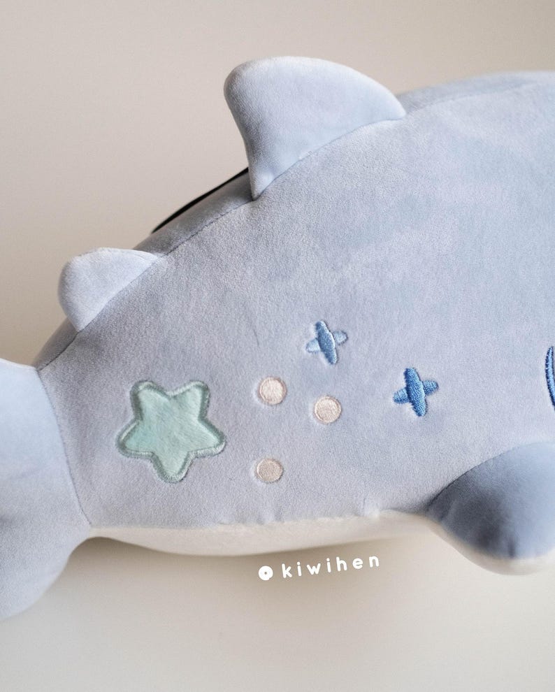 May include: A light blue plush whale with embroidered details, including a green star, white dots, and blue crosses. The whale has a white belly and a fin on its back. The text "kiwihen" is embroidered on the whale's belly.