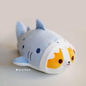 May include: A plush toy shaped like a blue shark with a white belly and a small orange and white dog peeking out of the shark's mouth.