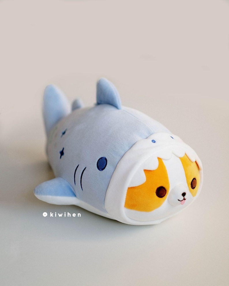 May include: A plush toy shaped like a blue shark with a white belly and a small orange and white dog peeking out of the shark's mouth.