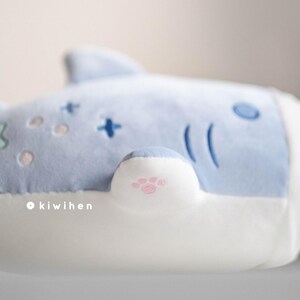 May include: A light blue plush toy shaped like a shark with a white belly. The shark has a pink paw print on its fin.