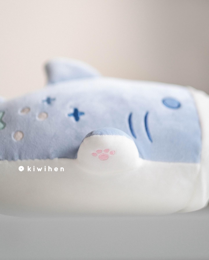 May include: A light blue plush toy shaped like a shark with a white belly. The shark has a pink paw print on its fin.