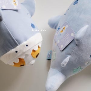 May include: Two plush blue shark toys with embroidered details. One shark has a white and yellow face with a toothy grin. The other shark has a white star and cloud pattern. Both sharks have a tag with the text "It's okay to be different, you're perfectly unique!"