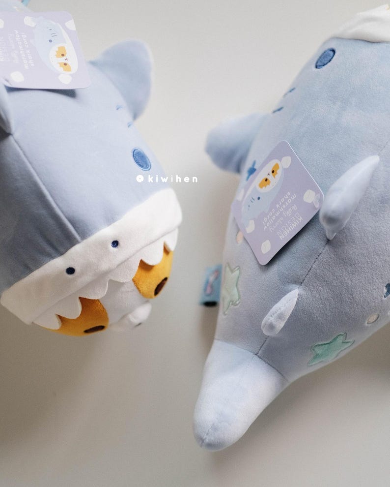 May include: Two plush blue shark toys with embroidered details. One shark has a white and yellow face with a toothy grin. The other shark has a white star and cloud pattern. Both sharks have a tag with the text "It's okay to be different, you're perfectly unique!"