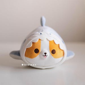 May include: A plush toy shaped like a shark with a cute cartoon face. The shark is white with blue fins and a yellow and white striped belly. The toy has big brown eyes, a pink nose, and a small mouth.