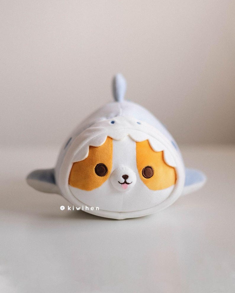 May include: A plush toy shaped like a shark with a cute cartoon face. The shark is white with blue fins and a yellow and white striped belly. The toy has big brown eyes, a pink nose, and a small mouth.