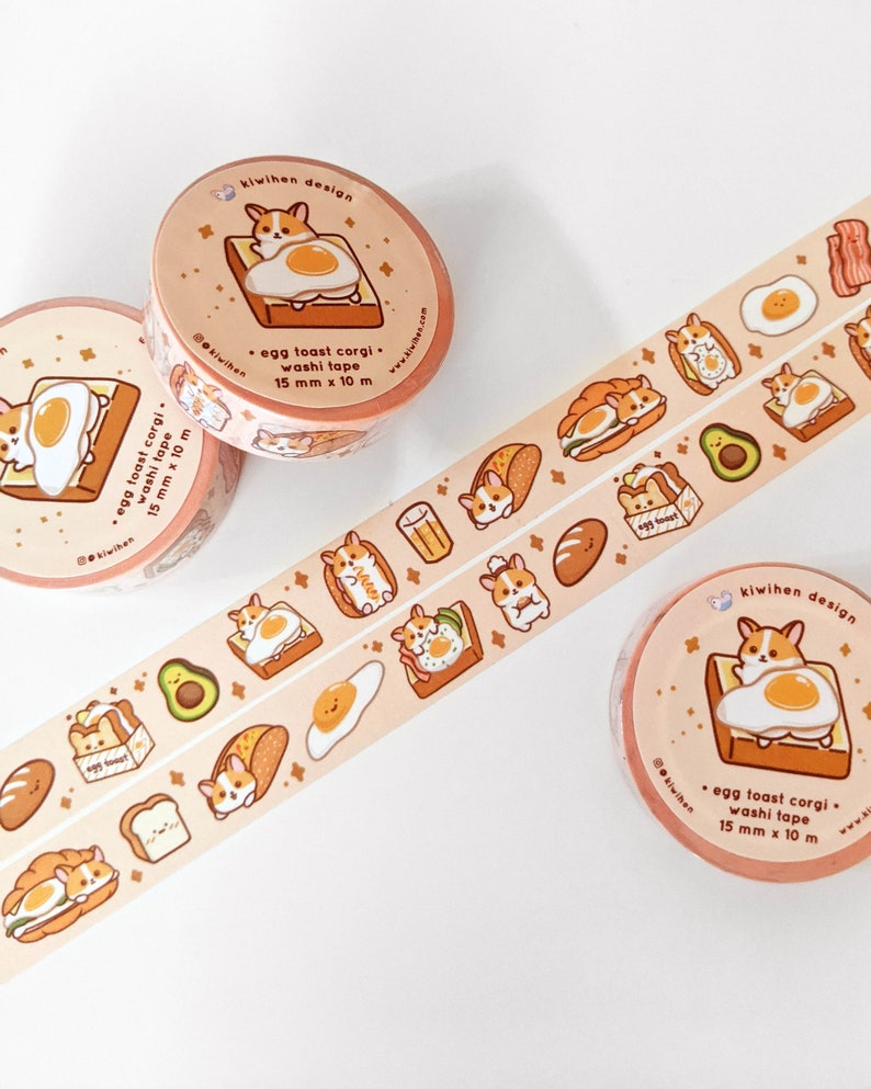 May include: Three rolls of washi tape featuring a cartoon corgi dog on a slice of toast with a fried egg. The tape is peach colored with a white background and has a repeating pattern of the corgi dog, toast, eggs, avocado, and other breakfast foods. The text on the tape reads "egg toast corgi. washi tape 15 mm x 10 m".