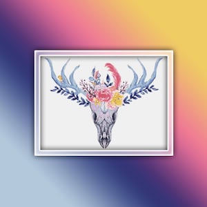 Puede incluir: Un cráneo de ciervo blanco con cuernos azules está adornado con una corona floral de flores rosas, amarillas y blancas. El cráneo está rodeado por un marco blanco.