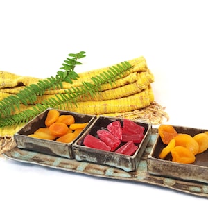 Peut inclure: Un plateau de service en céramique avec trois compartiments carrés contenant chacun des fruits secs différents. Le plateau est décoré d'un glaçage bleu et marron. Un tissu jaune et une fougère verte sont en arrière-plan. Les fruits secs sont orange et rouges.