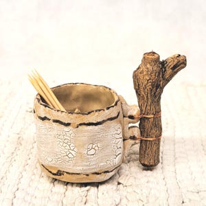 May include: A handcrafted ceramic toothpick holder with a rustic aesthetic. The off-white holder features a cracked texture and dark brown stripes. A natural wood handle is attached with copper wire, and several toothpicks are inside.
