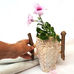 Puede incluir: Un jarrón de cerámica hecho a mano, de color blanquecino y con una textura trabajada. Palos de madera están fijados a los lados con alambre de cobre. El jarrón contiene una planta verde con flores rosas.
