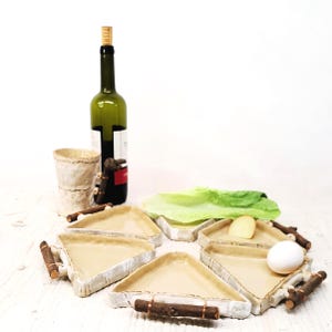Può includere: Una natura morta con una bottiglia di vino verde, una tazza di ceramica e sei piatti triangolari in ceramica con manici in legno. I piatti circondano una foglia di lattuga, un pezzo di formaggio e un uovo.