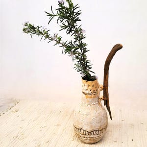 以下が含まれることがあります： ひび割れた仕上げが施された手作りのセラミック花瓶。装飾的な木の枝とローズマリーの小枝で飾られています。花瓶はクリーム色のベースに茶色のアクセントとユニークなハンドルが付いています。ローズマリーには小さな紫色の花が咲いています。