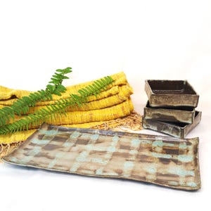 May include: A rectangular ceramic serving platter with a brown and blue chequered pattern. Three small square ceramic dishes are stacked on the right. A yellow cloth and fern are in the background.