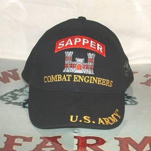 May include: A black baseball cap featuring an embroidered red arch with the white text SAPPER, a white and red castle emblem, and the yellow text COMBAT ENGINEERS. The brim displays the yellow embroidered text U.S. ARMY on the right side.