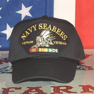 Puede incluir: Gorra negra con la inscripción "Navy Seabees Vietnam Veteran" bordada en oro y blanco. La gorra presenta un parche con un emblema de abeja y cintas militares. El fondo incluye una bandera americana.