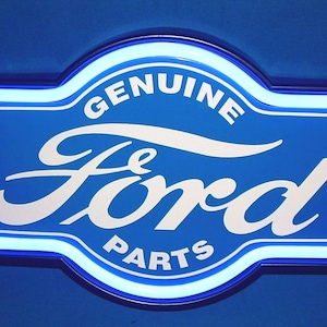 May include: A blue and white Ford Genuine Parts sign with a neon-style border. The sign features the Ford logo and the words "GENUINE PARTS" in white lettering. The sign is rectangular with rounded edges.
