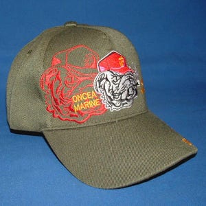 May include: Olive green baseball cap with embroidered design. Features a red and white bulldog wearing a red hat, and the words "ONCEA MARINE" in red. The cap has a curved brim.