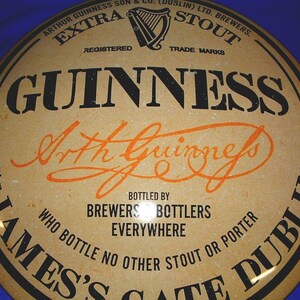 K&ouml;nnte beinhalten: Rundes Vintage-Guinness-Stout-Schild mit schwarzem und orangefarbenem Text auf beigem Hintergrund. Das Schild zeigt das Guinness-Logo, die Worte "EXTRA STOUT" und "Arthur Guinness" in Schrift. Es lautet "Abgef&uuml;llt von Brewers Bottlers Everywhere".