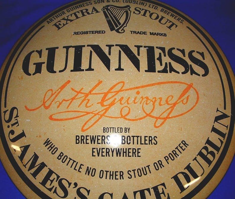 K&ouml;nnte beinhalten: Rundes Vintage-Guinness-Stout-Schild mit schwarzem und orangefarbenem Text auf beigem Hintergrund. Das Schild zeigt das Guinness-Logo, die Worte "EXTRA STOUT" und "Arthur Guinness" in Schrift. Es lautet "Abgef&uuml;llt von Brewers Bottlers Everywhere".