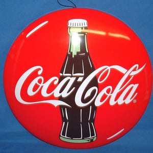 Könnte beinhalten: Rundes, rotes Coca-Cola-Schild mit einem klassischen Flaschendesign in der Mitte. Das Coca-Cola-Logo ist weiß, mit einer schwarzen Flaschenkontur und einem weißen Verschluss. Oben ist eine schwarze Schnur zum Aufhängen angebracht.