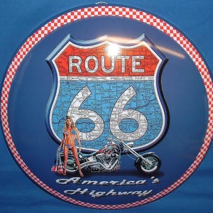 Puede incluir: Un letrero redondo azul de la Ruta 66 con un borde a cuadros rojo y blanco. El letrero presenta un mapa de la ruta, las palabras "Route 66" y una motocicleta. Las palabras "America's Highway" están en la parte inferior. Aproximadamente 30 cm de diámetro.