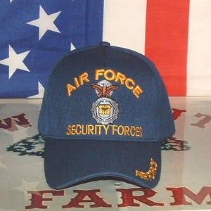 May include: A navy blue baseball cap with the words "AIR FORCE SECURITY FORCES" embroidered in gold. The cap features a patch with an eagle and shield design. The background includes an American flag.