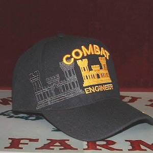 May include: A black baseball cap featuring an embroidered gold castle emblem and the text COMBAT ENGINEER in gold lettering. To the left of the gold castle is a secondary, outlined castle design embroidered in a dark grey thread color.