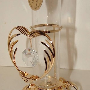 May include: A gold-toned crystal holder with a clear glass tube. The holder features a heart-shaped design with a dangling crystal and a floral base. The text on the tag reads "The Original Crystal Delight".