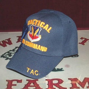 May include: A navy blue baseball cap with the words "TACTICAL AIR COMMAND" embroidered in gold thread. The cap features a patch with a winged emblem and the letters "TAC" embroidered on the brim.