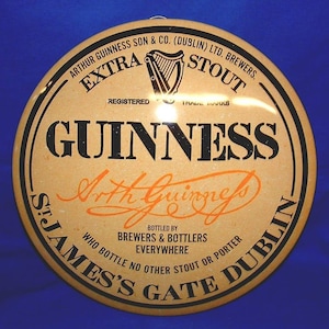 K&ouml;nnte beinhalten: Rundes Guinness Stout-Schild mit beigem Hintergrund und schwarzem und orangefarbenem Text. Das Schild zeigt das Guinness-Logo, die Worte "Extra Stout" und "St. James's Gate Dublin."