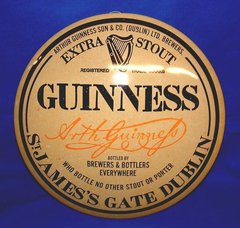 K&ouml;nnte beinhalten: Rundes Guinness Stout-Schild mit beigem Hintergrund und schwarzem und orangefarbenem Text. Das Schild zeigt das Guinness-Logo, die Worte "Extra Stout" und "St. James's Gate Dublin."