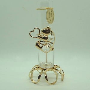 May include: A clear glass bud vase with a gold-colored metal teddy bear holding a heart. The bear has crystal accents. The vase sits on a gold-colored metal base. A tag is attached to the vase.