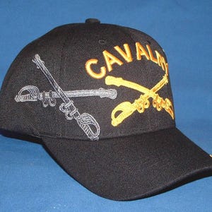 May include: A black baseball cap featuring embroidered crossed sabers on the front. One saber is stitched in silver thread, while the other is in gold. The word CAVALRY is embroidered in gold thread above the crossed sabers on the front panel.