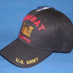 May include: A black baseball cap featuring the embroidered red text COMBAT and ENGINEER with a gold castle insignia in the center. The brim displays the yellow embroidered text U.S. ARMY. A small white tag is attached to the top of the crown.