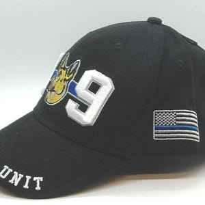 May include: Black baseball cap with white embroidered "K-9 UNIT" text on the brim. The cap features a K-9 dog graphic and the number "9" in white. A small American flag patch with a blue line is on the side.