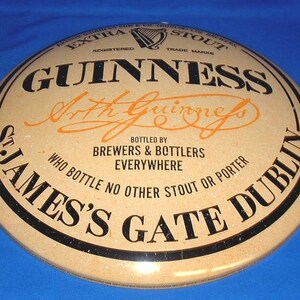 K&ouml;nnte beinhalten: Ein rundes Guinness-Schild mit beigem Hintergrund und schwarzer und orangefarbener Schrift. Das Schild zeigt das Guinness-Logo, den Namen "Arthur Guinness" und den Text "St. James's Gate Dublin" und "Extra Stout".