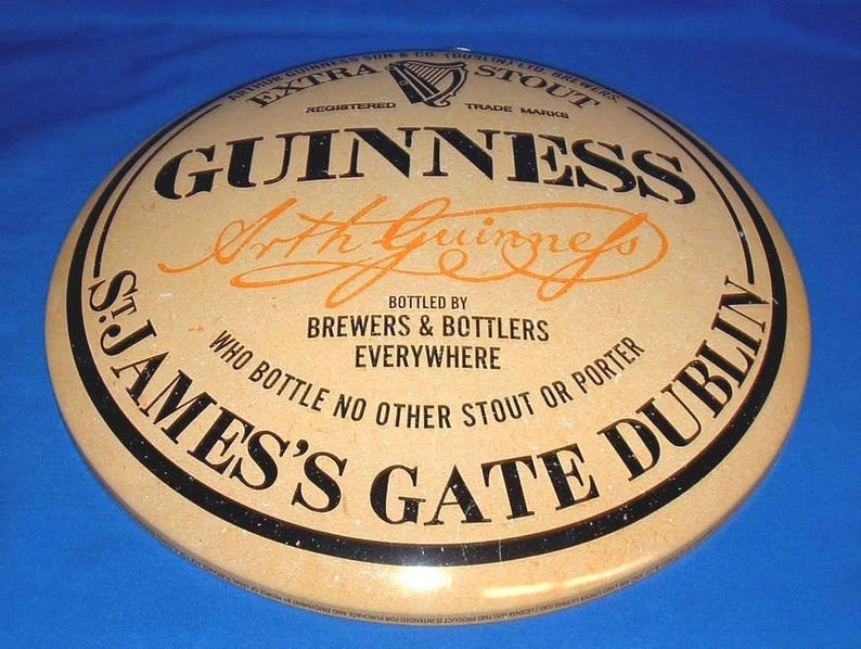 K&ouml;nnte beinhalten: Ein rundes Guinness-Schild mit beigem Hintergrund und schwarzer und orangefarbener Schrift. Das Schild zeigt das Guinness-Logo, den Namen "Arthur Guinness" und den Text "St. James's Gate Dublin" und "Extra Stout".