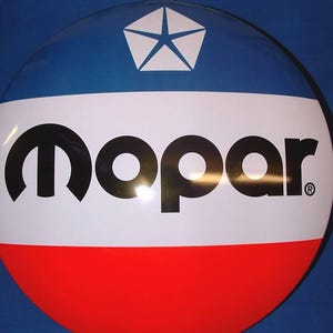 Pode incluir: Uma placa de metal circular com um design tricolor horizontal em azul, branco e vermelho. A seção central branca exibe o texto preto Mopar com um símbolo de marca registrada. Um logotipo pentastar branco está centralizado na seção azul.