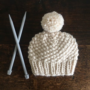 Peut inclure: Bonnet en tricot crème avec un pompon sur le dessus et deux aiguilles à tricoter grises croisées devant. Le bonnet a un motif de point texturé et un bord côtelé. Les aiguilles sont de taille 12.