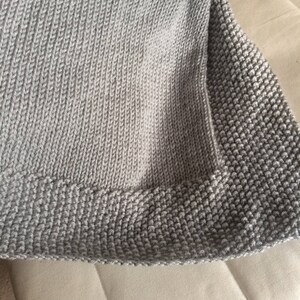 May include: A gray knitted throw blanket with a textured border. The blanket is folded and draped over a light beige surface.