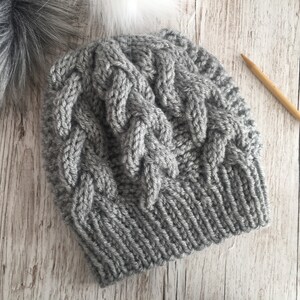 May include: A gray knitted beanie with a cable knit pattern. The beanie is made with a soft yarn and has a simple, classic design.