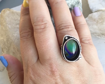 Puede incluir: Un anillo de plata con una piedra grande de forma ovalada iridiscente engastada en un engaste de plata. La piedra es de color verde oscuro con toques de azul y morado.