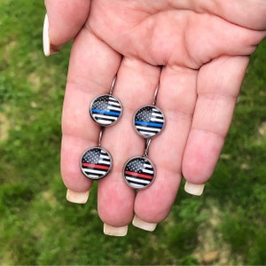 May include: A pair of silver dangle earrings with round glass charms featuring the American flag with a thin red, white, and blue stripe across the middle.