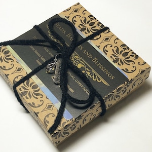 May include: A small brown box with a black ribbon tied around it. The box has a gold and black patterned design. The ribbon has a silver charm attached to it that says "EVER". The box has a label that says "BEADS, BA ES AND BLESSINGS RITUAL, GYPSY SOUL oo.com".