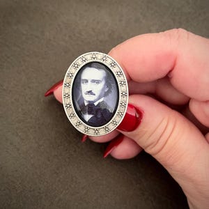 May include: Oval-shaped silver ring featuring a portrait of Edgar Allan Poe. The portrait is in black and white, set within a decorative frame. The ring is held against a brown background.