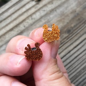 May include: A pair of gold glitter turkey stud earrings. The earrings are small and have a simple design.