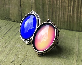 Puede incluir: Dos anillos de humor con engastes ovalados. El anillo de la izquierda tiene una piedra azul y el anillo de la derecha tiene una piedra rosa y amarilla.