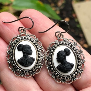 May include: A pair of silver earrings with a cameo design featuring a black silhouette of a woman's head with a bun. The cameo is set in a white oval frame with a silver filigree border.