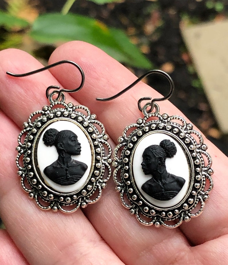 May include: A pair of silver earrings with a cameo design featuring a black silhouette of a woman's head with a bun. The cameo is set in a white oval frame with a silver filigree border.